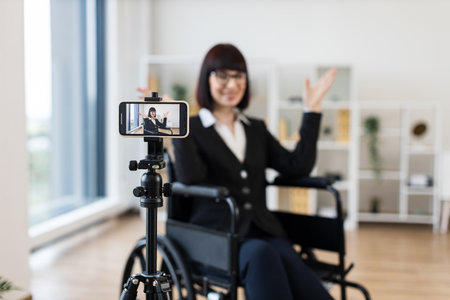 Caucasian woman in business attire sits in wheelchair recording informative business blog on smartphone. Modern interior, professional appearance, creating content for business communication.の写真素材