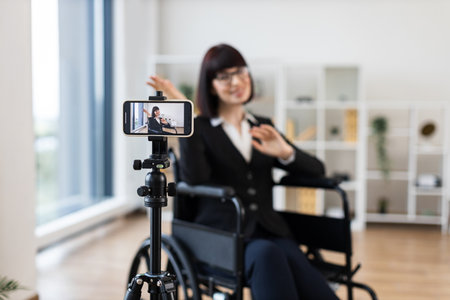 Caucasian woman in business attire sits in wheelchair recording informative business blog on smartphone. Modern interior, professional appearance, creating content for business communication.の写真素材