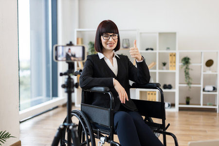 Caucasian woman in wheelchair wearing business attire blogging on camera in bright office space. Promotes concepts of inclusive business, motivation, and professionalism.の写真素材