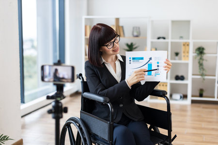 Caucasian woman in wheelchair holding chart showing business data while blogging on cameraの写真素材