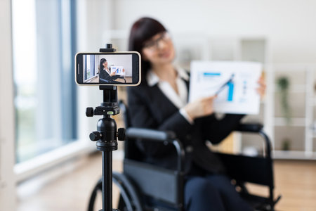 Caucasian woman in wheelchair blogs on camera about business using charts and graphs. Indoor setting with focus on professional blogging and business concepts.の写真素材