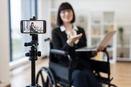 Caucasian adult woman sitting in wheelchair recording business presentation with smartphone and...
