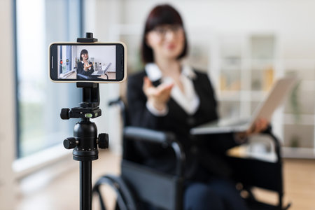 Caucasian woman in smart suit sitting in wheelchair in office during video blogging session about business practices using smartphone on tripod, promoting communication, inclusivityの写真素材