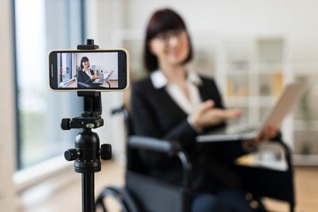 Caucasian woman in smart suit sitting in wheelchair in office during video blogging session about business practices using smartphone on tripod, promoting communication, inclusivityの写真素材