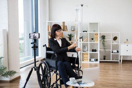 Caucasian woman, wearing formal attire, sits in wheelchair and records blog using camera. Indoor setting emphasizes profession and resilience, highlighting adaptability during treatmentの写真素材
