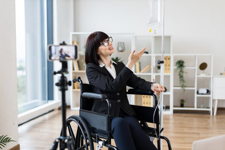 Caucasian woman in wheelchair with intravenous drip recording blog about handling treatment with work. She conveys empowerment and adaptability in modern life, highlighting professional and balance.の写真素材