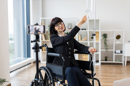 Caucasian woman in wheelchair with intravenous drip records video about combining work and health in modern professional lifestyle. Represents determination, adaptabilityの写真素材