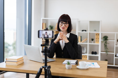 Caucasian young woman wearing suit sitting at office desk recording professional video with smartphone. Scene suggests concepts of communication, workplace modernity, online engagement, and knowledge.の写真素材