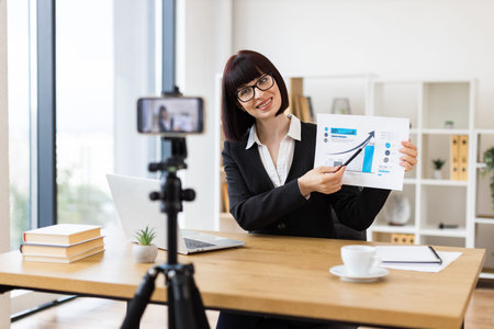 Caucasian adult woman with glasses at desk recording blog about business analysis and growth. Captures concepts like communication business trends remote work education and professional development.の写真素材