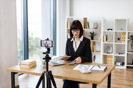 Caucasian woman, adult, wearing formal attire sitting at desk in office environment engaging in blog creation about investments, financial literacy, and income insights while holding dollar cash.の写真素材