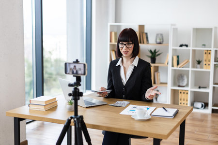 Caucasian woman in office recording vlogging session discussing financial literacy, investments, income. Holding dollar bills, explaining strategies. Modern equipment setup, professionally dressed.の写真素材
