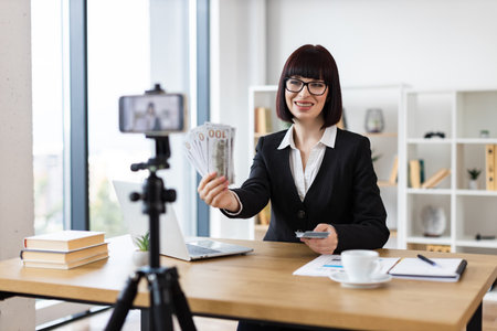 Caucasian woman, mid-thirties, presenting financial concepts on video, wearing formal attire, holding cash, discussing investments and income strategies, set in modern office with laptopの写真素材