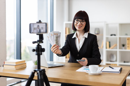 Caucasian woman in office presenting financial concepts on video. Discussing income sources, investments, and effective money management while holding cash.の写真素材