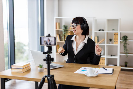 Caucasian woman wearing glasses and headphones using laptop in office for work. Office interior includes shelves, books, plants, and phone on tripod. Concept of remote work, modern technology.の写真素材