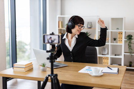 Caucasian woman wearing suit sitting at desk with headphones recording blog relaxing to music. Scene blends professional work with personal enjoyment in a modern office setting highlighting balance.の写真素材