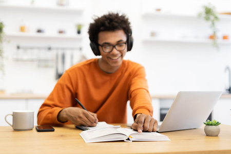 A young man wearing headphones smiles while studying at a table with a laptop and notebook.