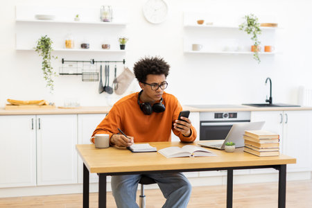 A young man with glasses is working at a table in his kitchen, looking at his phone with a concerned expression.の写真素材