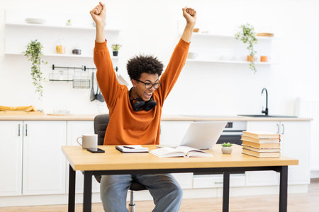 A young man celebrates a win while working on his laptop at home, arms raised in triumph.の写真素材