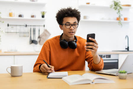 A young man with glasses is using his phone while working at a table with a laptop and notebook.の写真素材