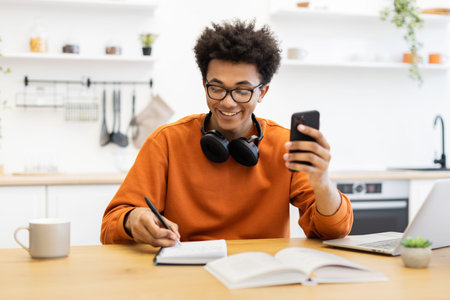 A young man with glasses smiles while looking at his phone and writing in a notebook at a table with...