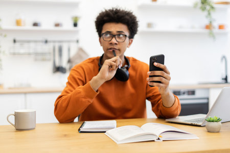 A young man in glasses contemplates while holding a phone and pen, with a book and laptop on the table.の写真素材