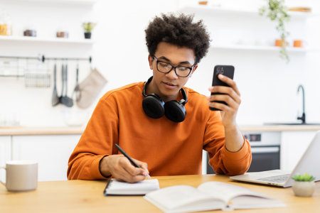 A young man with glasses is working at a table, using his phone and writing in a notebook.の写真素材