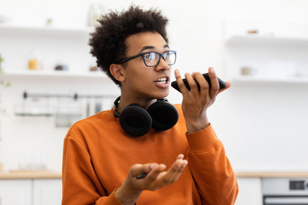 A young man with glasses speaks into his smartphone, likely using a voice assistant or making a call, with headphones around his neck.の写真素材