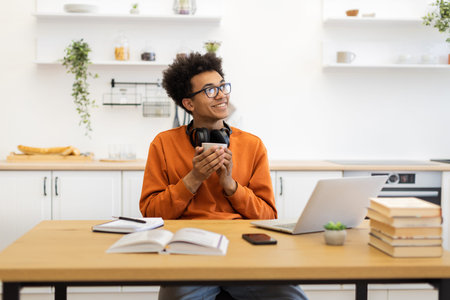 A young man with glasses smiles while holding a cup of coffee in his kitchen, with a laptop and...