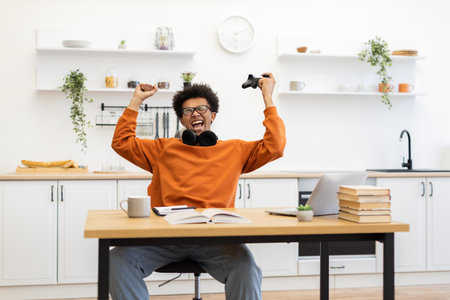 A young man celebrates a win while gaming in his kitchen. He is holding a game controller and is very excited.の写真素材
