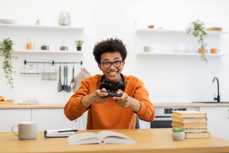 A young man with glasses is enthusiastically playing a video game in his kitchen, holding a...