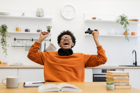 A young man with glasses celebrates a win while playing video games at home, holding a controller in the air.の写真素材