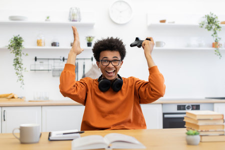 A young man with glasses celebrates a gaming win, holding a controller in his raised hand with joy.の写真素材