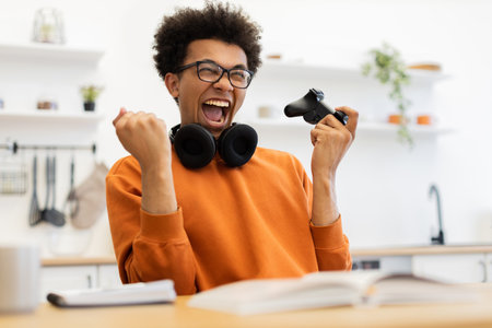 A young man with glasses celebrates a win while playing a video game, holding a controller.の写真素材