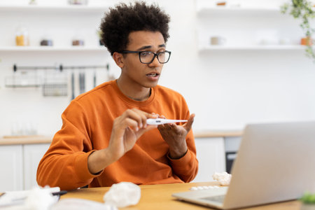 A young man with a thermometer is on a video call, possibly discussing his health while working from...