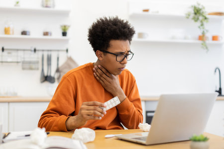 A young man with glasses holds medicine while experiencing a sore throat, possibly consulting a doctor online.の写真素材