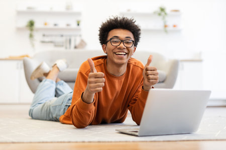 A young man with glasses smiles and gives a thumbs up while using a laptop at home.の写真素材