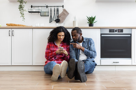 A couple sits on the floor in their kitchen, looking at a phone together. The woman is holding the phone, and the man is pointing at it.の写真素材