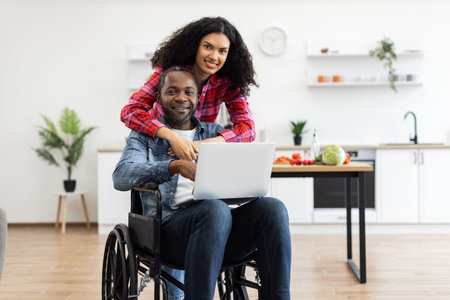 A smiling couple, one in a wheelchair, poses together in a modern kitchen with a laptop.の写真素材