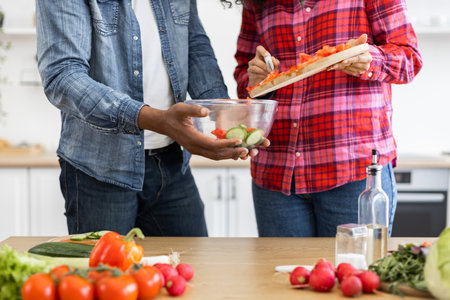 A couple is shown in a kitchen, preparing a salad with fresh vegetables. One person is adding chopped tomatoes to a bowl.の写真素材
