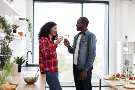 A diverse couple enjoys a moment together, toasting with wine glasses in a bright, contemporary kitchen setting.の写真素材