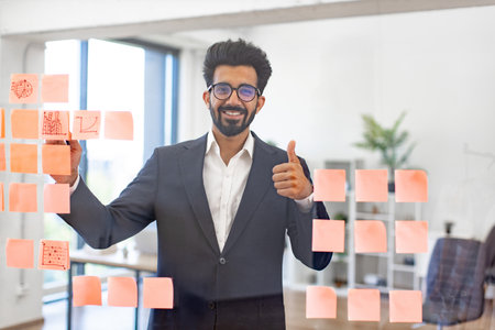 An Indian businessman smiles while working on a project with sticky notes, showing a thumbs up gestureの写真素材