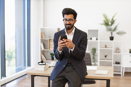 An Indian businessman in a suit smiles while using his smartphone in a modern office setting