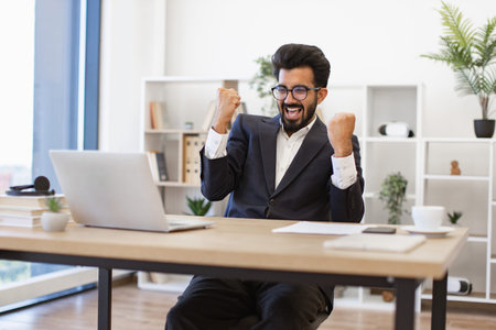 An excited businessman celebrates a win in his office, looking at his laptop screen
