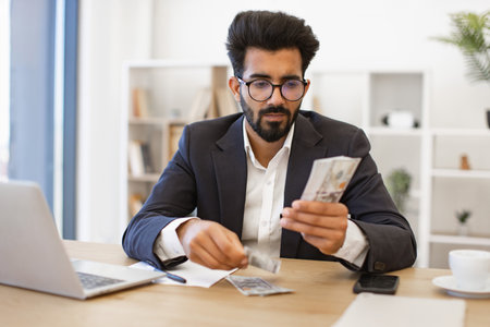 A businessman is carefully counting a stack of dollar bills at his desk, possibly for financial planning or investment purposesの写真素材