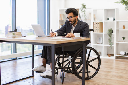 An Indian businessman in a wheelchair is working in his modern office, focused on his tasksの写真素材