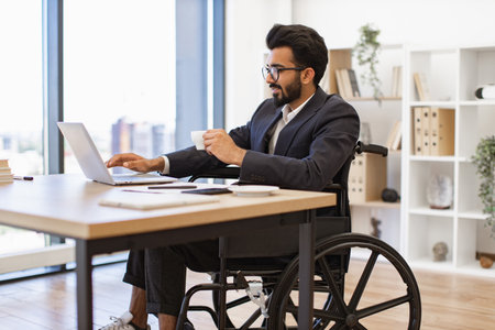 A businessman in a wheelchair works on a laptop while holding a cup of coffee in a modern office settingの写真素材