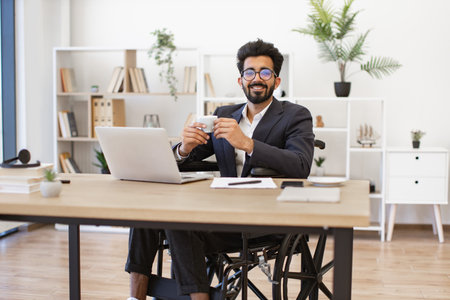A smiling businessman in a wheelchair enjoys a cup of coffee while working at his deskの写真素材