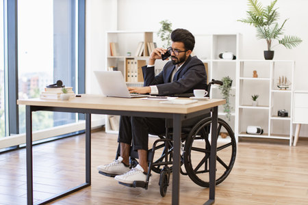 A businessman in a wheelchair works in his office, using a laptop and talking on the phoneの写真素材
