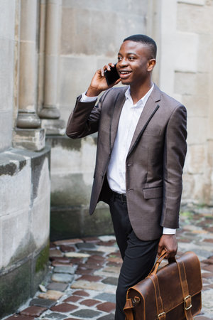 A smiling Black man in a suit walks down a cobblestone street while talking on his smartphone and carrying a leather briefcaseの写真素材