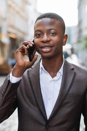 A young Black man in a suit and white shirt is talking on his smartphone while standing outdoors on...